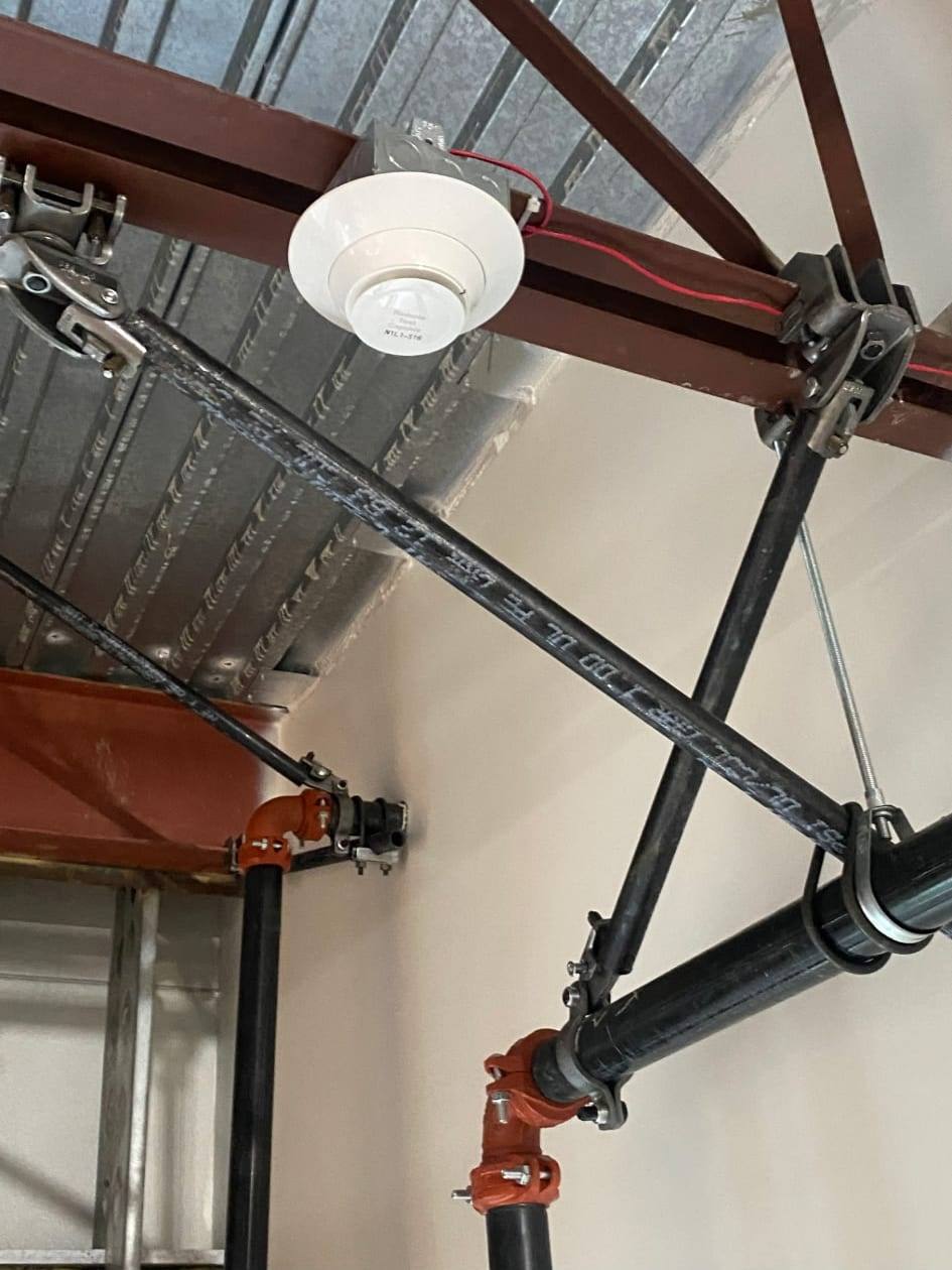 Fire sprinkler branch line supported by seismic sway bracing against a metal deck, with a ceiling-mounted heat detector alongside