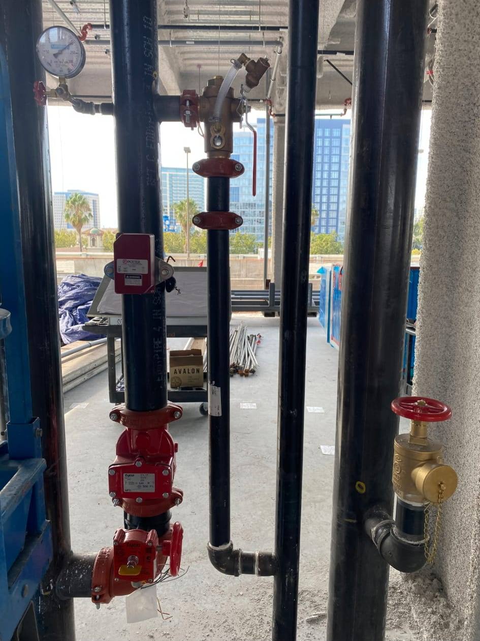 Fire standpipe riser with a pressure gauge, flow switch, control valve and brass hose valve installed on an open floor of a building under construction, city skyline behind